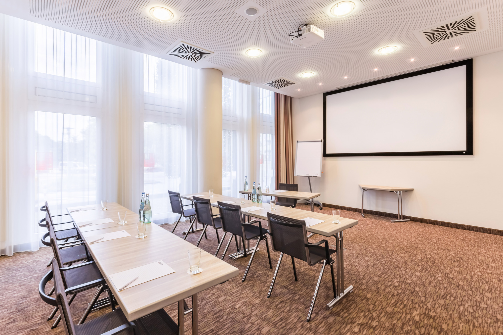 Kleiner Tagungsraum mit zwei Tischreihen, Beamer an der Decke und einem Tisch vor Flipchart und Leinwand